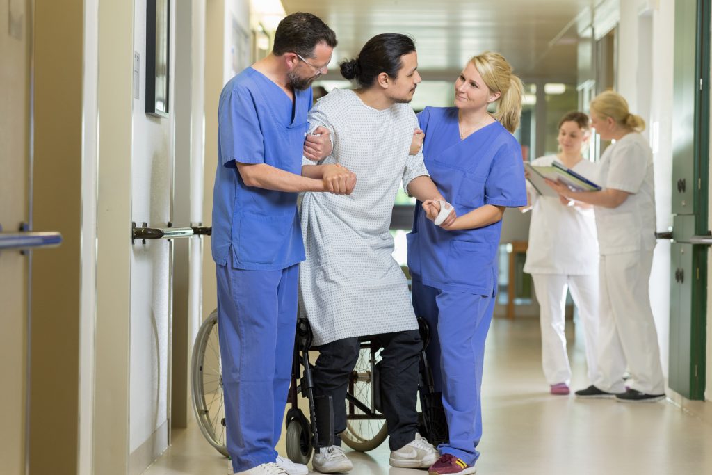 Zwei Pflegefachkräfte helfen einem Patienten aus seinem Rollstuhl