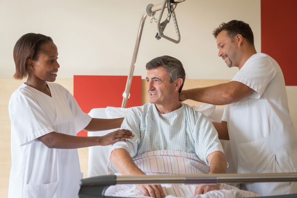 Zwei Pflegekräfte in der Ausbildung helfen einem Herren beim Aufstehen aus dem Krankenbett